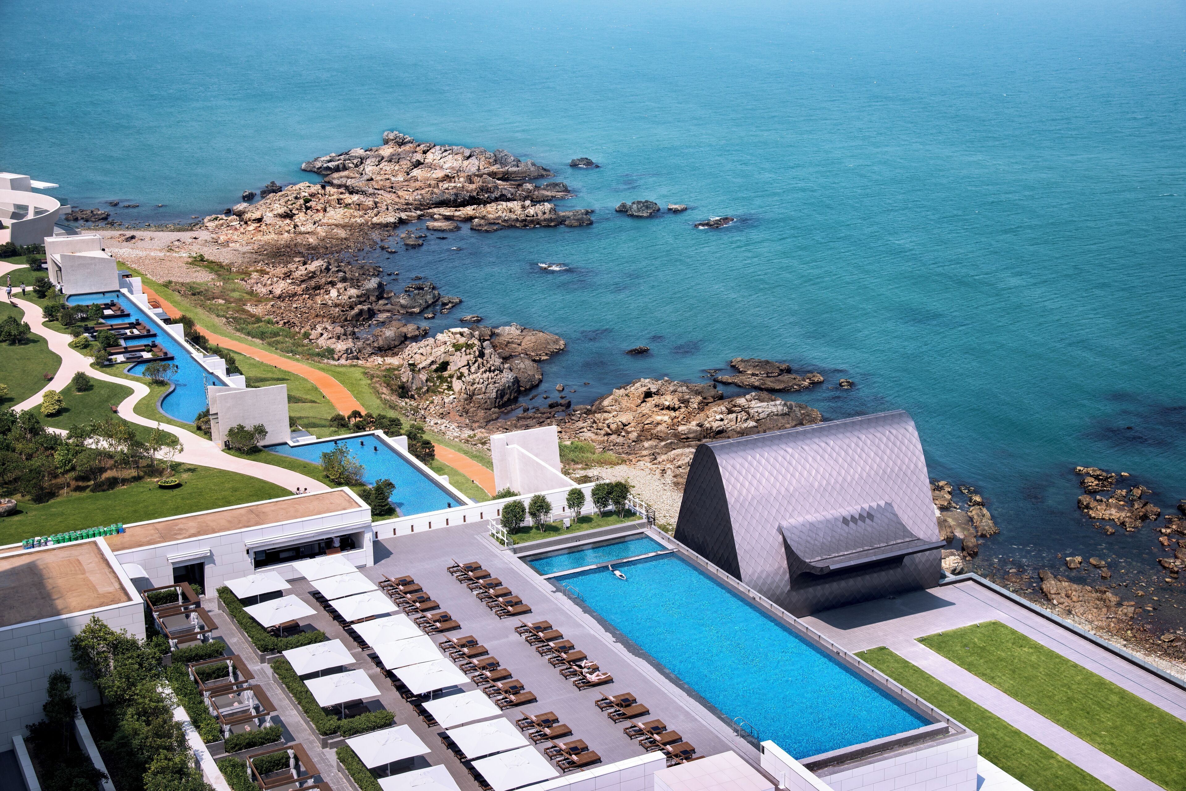 Ein Resort mit großem Pool, felsiger Küste und einem modernen Gebäude mit außergewöhnlichem Dachdesign.