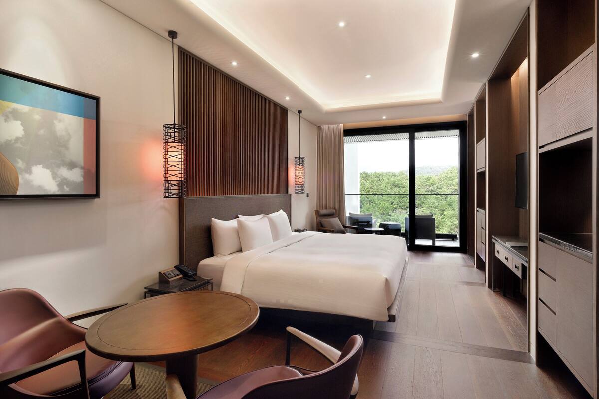 Ein modernes Hotelzimmer mit einem großen Bett, einem Schreibtisch, einem Sessel, einem Fernseher und einem Balkon mit Blick auf Grünflächen.