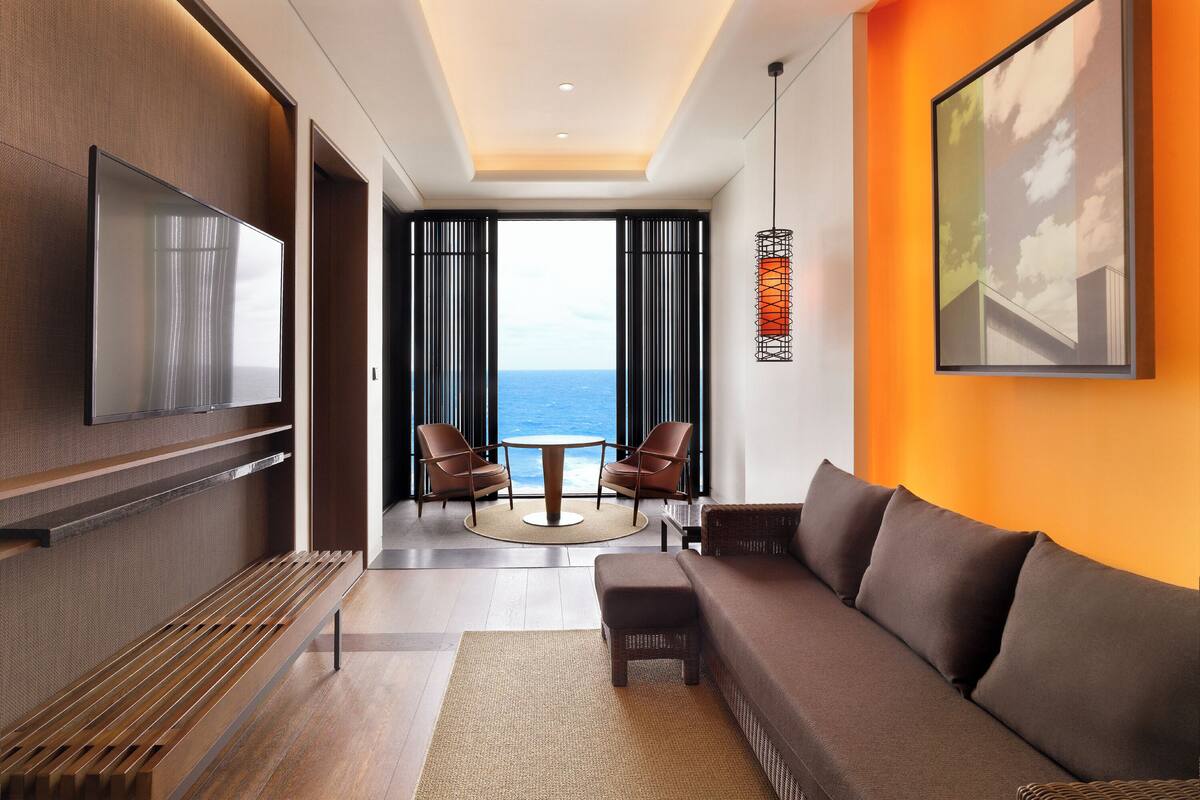 Ein modernes Hotelzimmer mit einer Couch, einem Fernseher, einem Essbereich mit Stühlen und Blick aufs Meer.