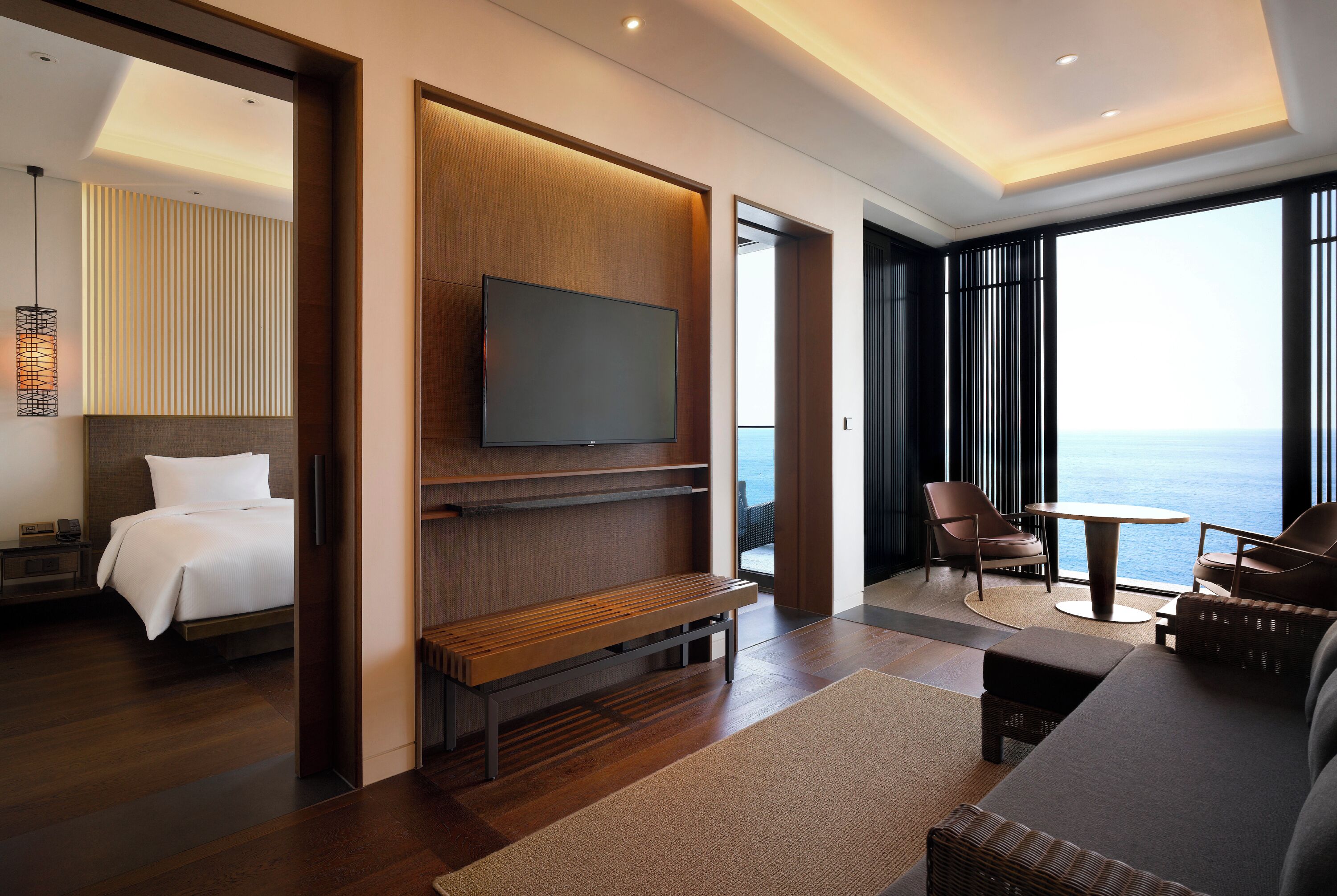 A modern hotel room with a bed, a TV, a seating area with a view of the sea, and a balcony.