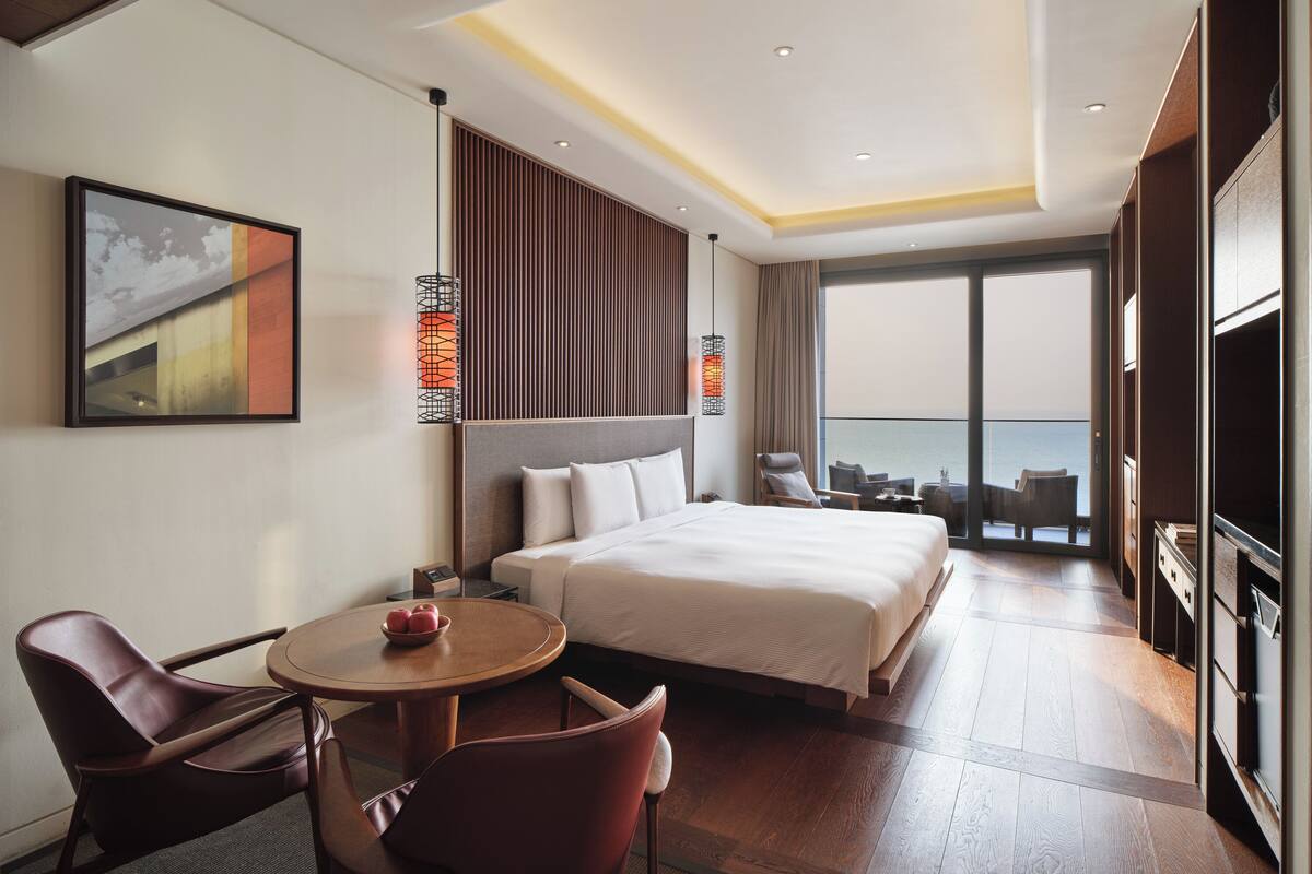 Ein Hotelzimmer mit einem großen Bett, einem Schreibtisch mit Stühlen, einem Esstisch und einem Balkon mit Meerblick.