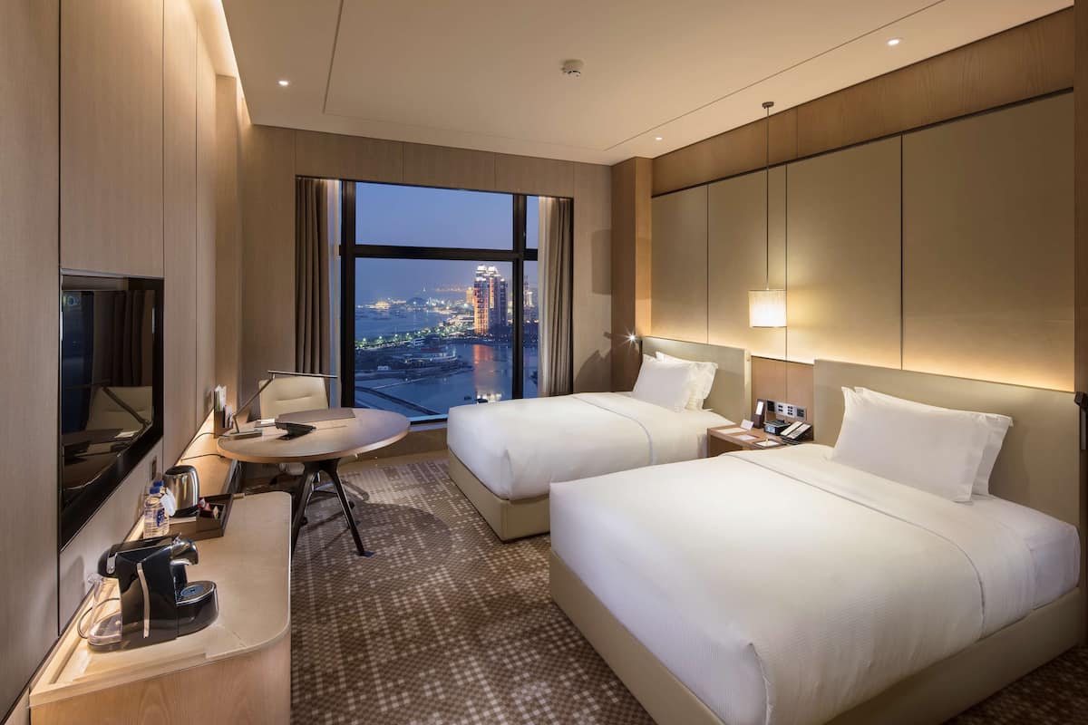Une chambre d’hôtel avec deux lits, un bureau, une chaise, une télévision et une grande fenêtre donnant sur la ville.