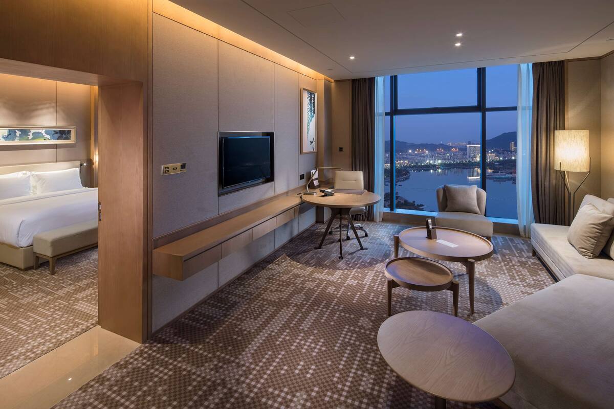 Une chambre d’hôtel moderne équipée d’un lit, d’une télévision, d’un coin salon avec une table et des chaises, et d’une grande fenêtre donnant sur la ville.