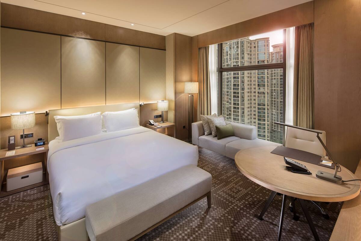 Une chambre d’hôtel avec un grand lit, un bureau avec une chaise, un canapé, une lampe et une vue sur le paysage urbain.