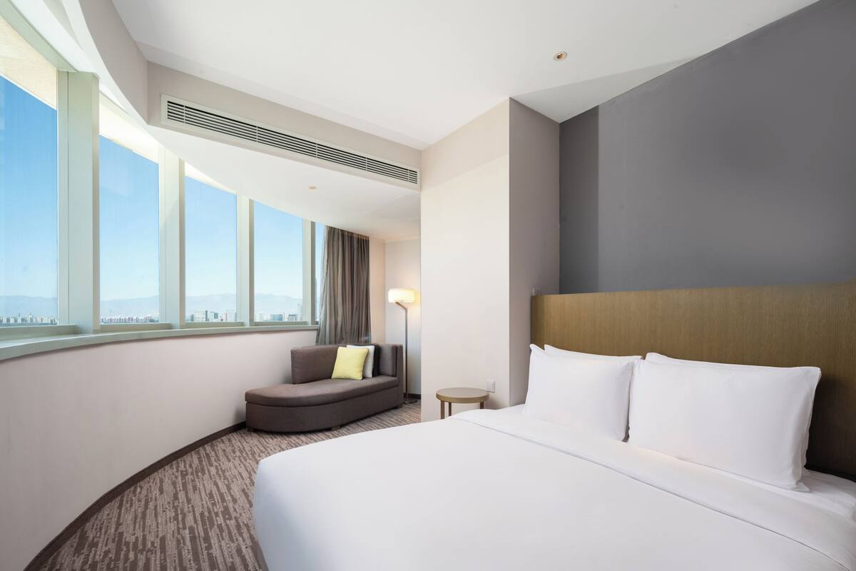 A hotel room with a large bed, a sofa, a small table, and a window with a city view.