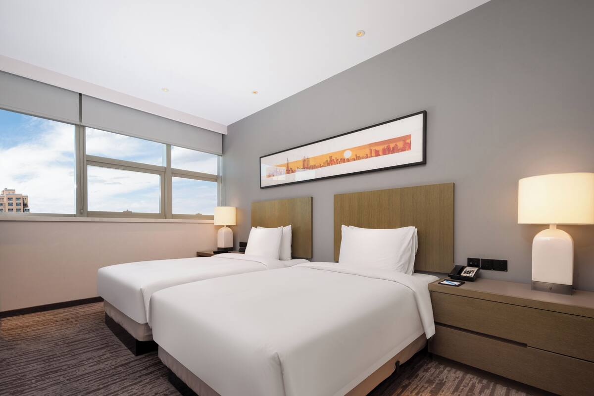 A hotel room with two beds, a cityscape view, a framed picture, and a nightstand with a lamp.