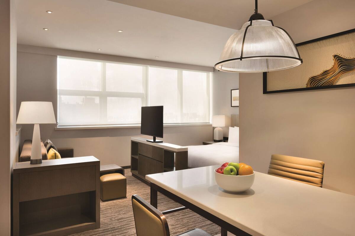 A hotel room with a bed, a desk, a dining table with fruit, a TV, and a large window.