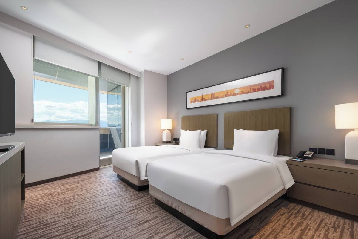 A hotel room with two beds, a large window, a painting, and a desk with a lamp.
