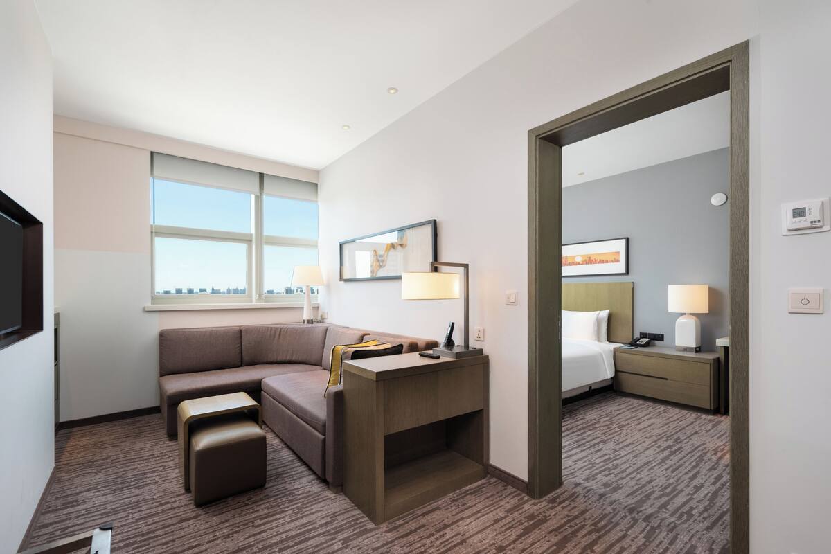 A hotel room with a sofa, a desk, a lamp, a bedside table, a mirror, and a window.