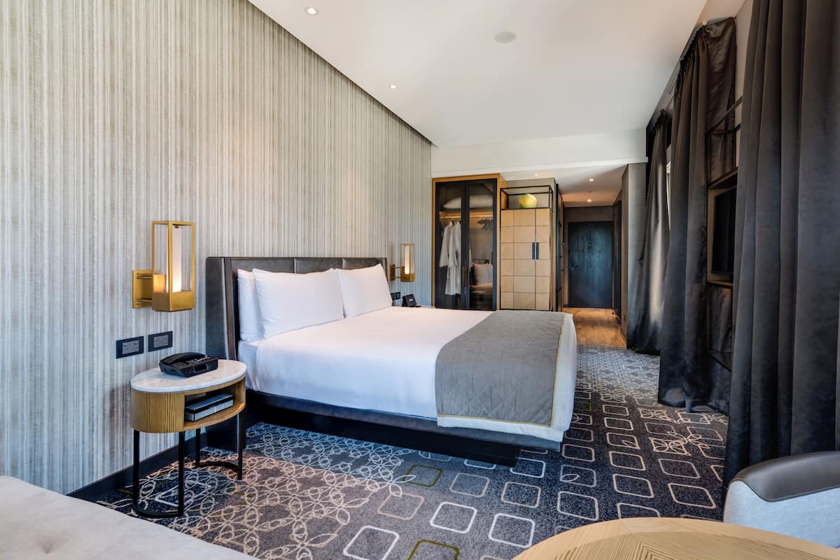 A modern hotel room with a large bed, a bedside table with a lamp, a wardrobe, and a patterned carpet.