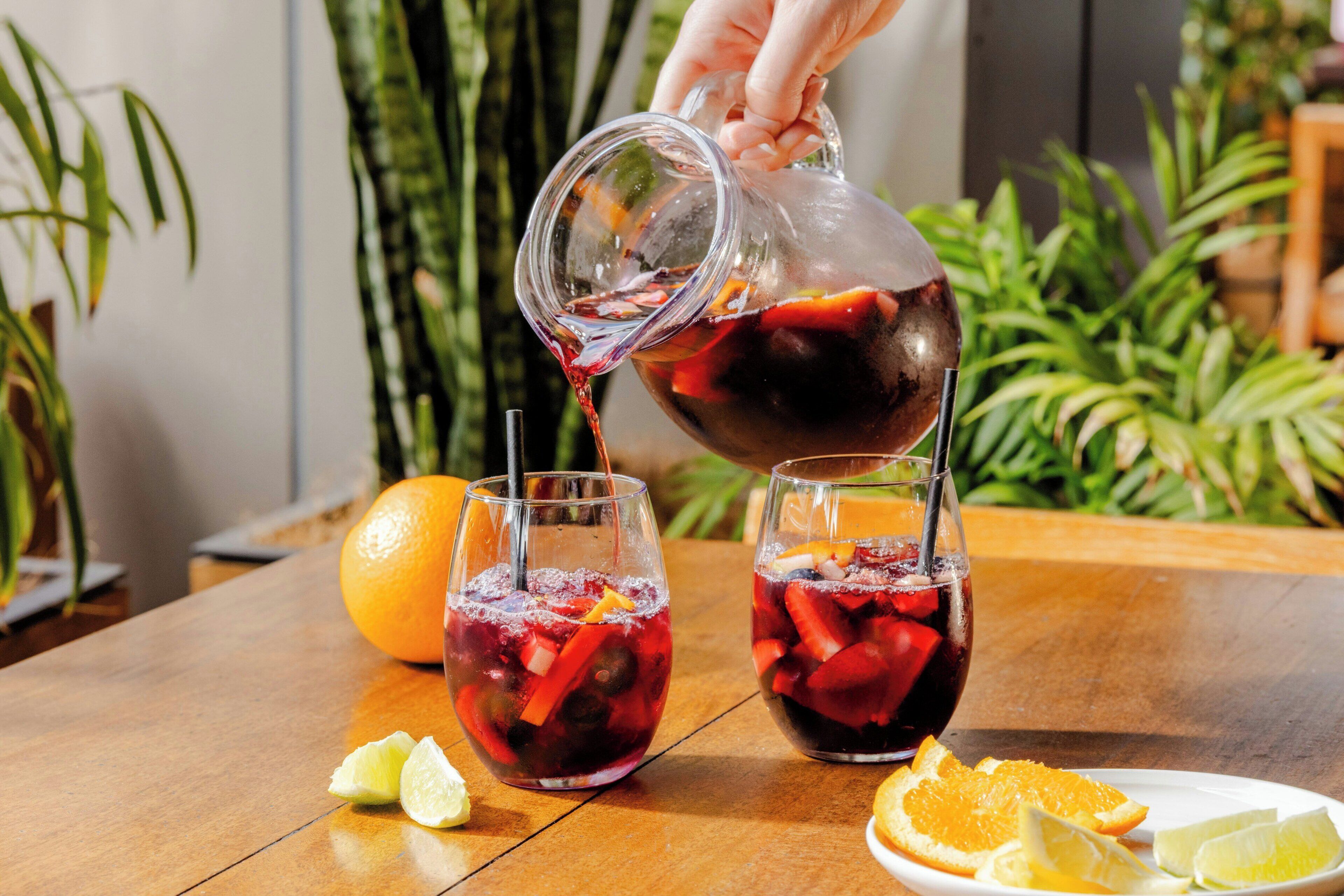 A hand pouring a drink into a glass with ice and fruit, with a lime wedge and orange slices on the table.