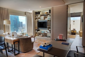 A modern hotel room with a large window, a flat-screen TV, a bookshelf, and a desk with a lamp.