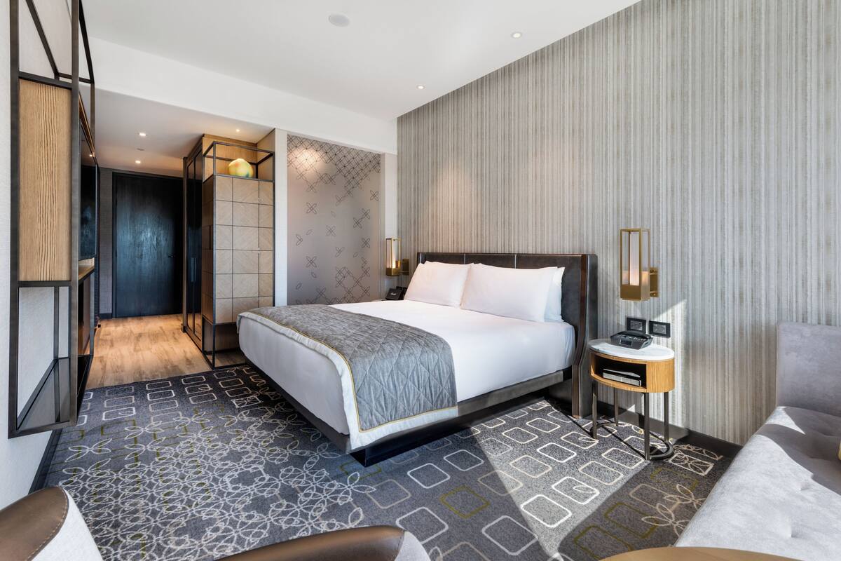 A modern hotel room with a large bed, a bedside table with a lamp, a small side table, and a patterned rug.