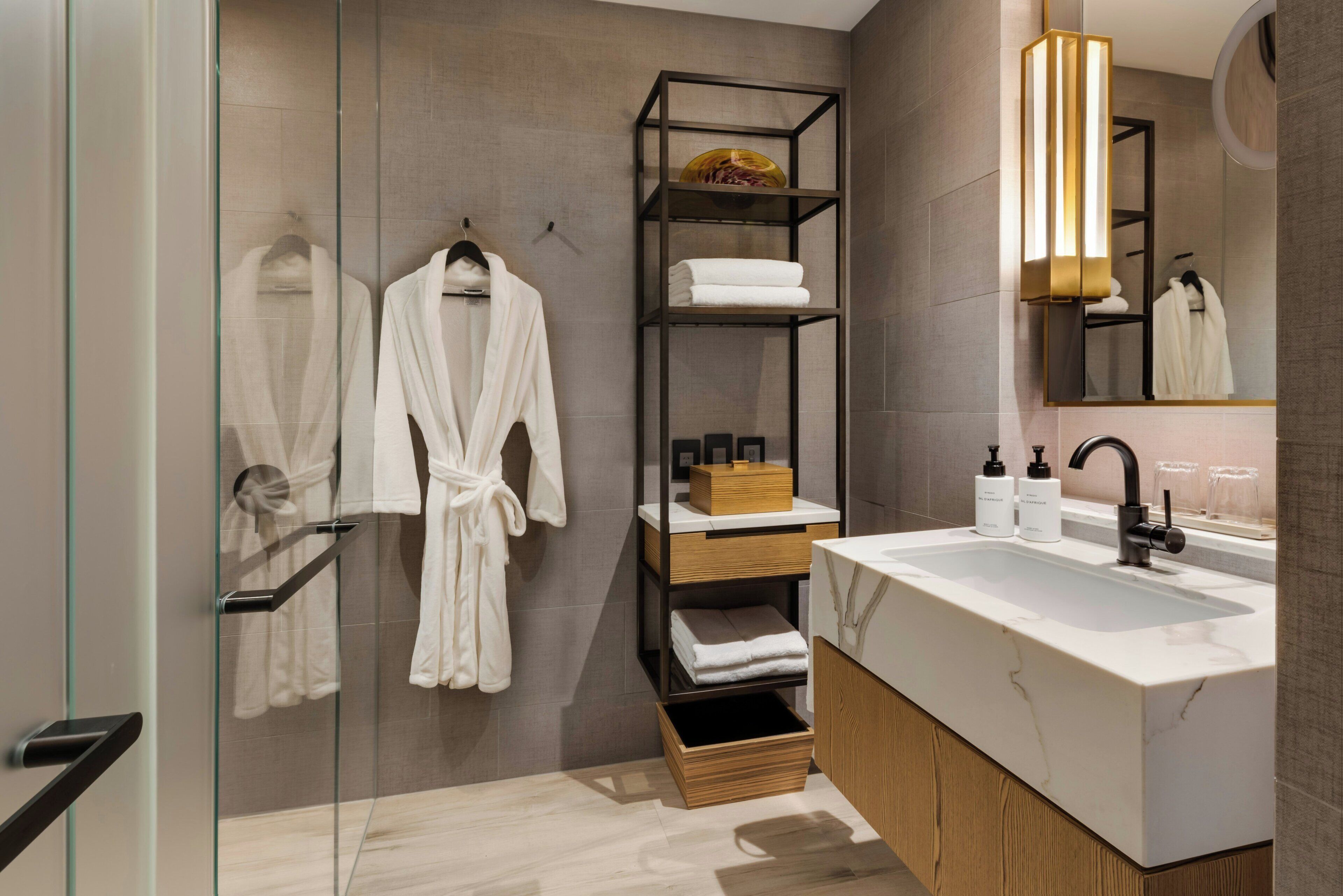 Un baño moderno con cabina de ducha de vidrio, tocador de madera con encimera de mármol y estantería de metal negro con toallas y artículos de aseo.