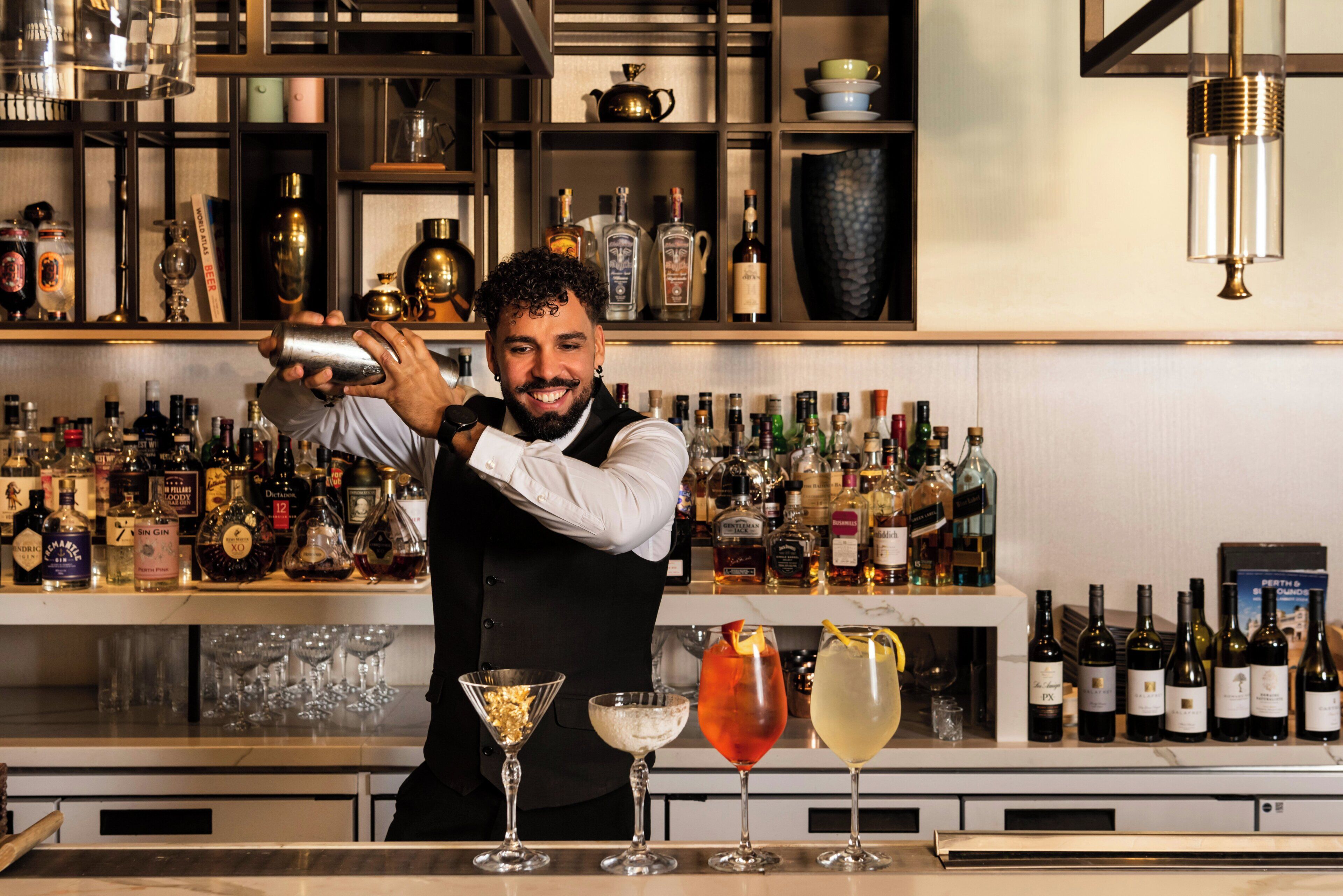 Un barman prépare un cocktail derrière un comptoir, avec différentes bouteilles et différents verres à portée de main.