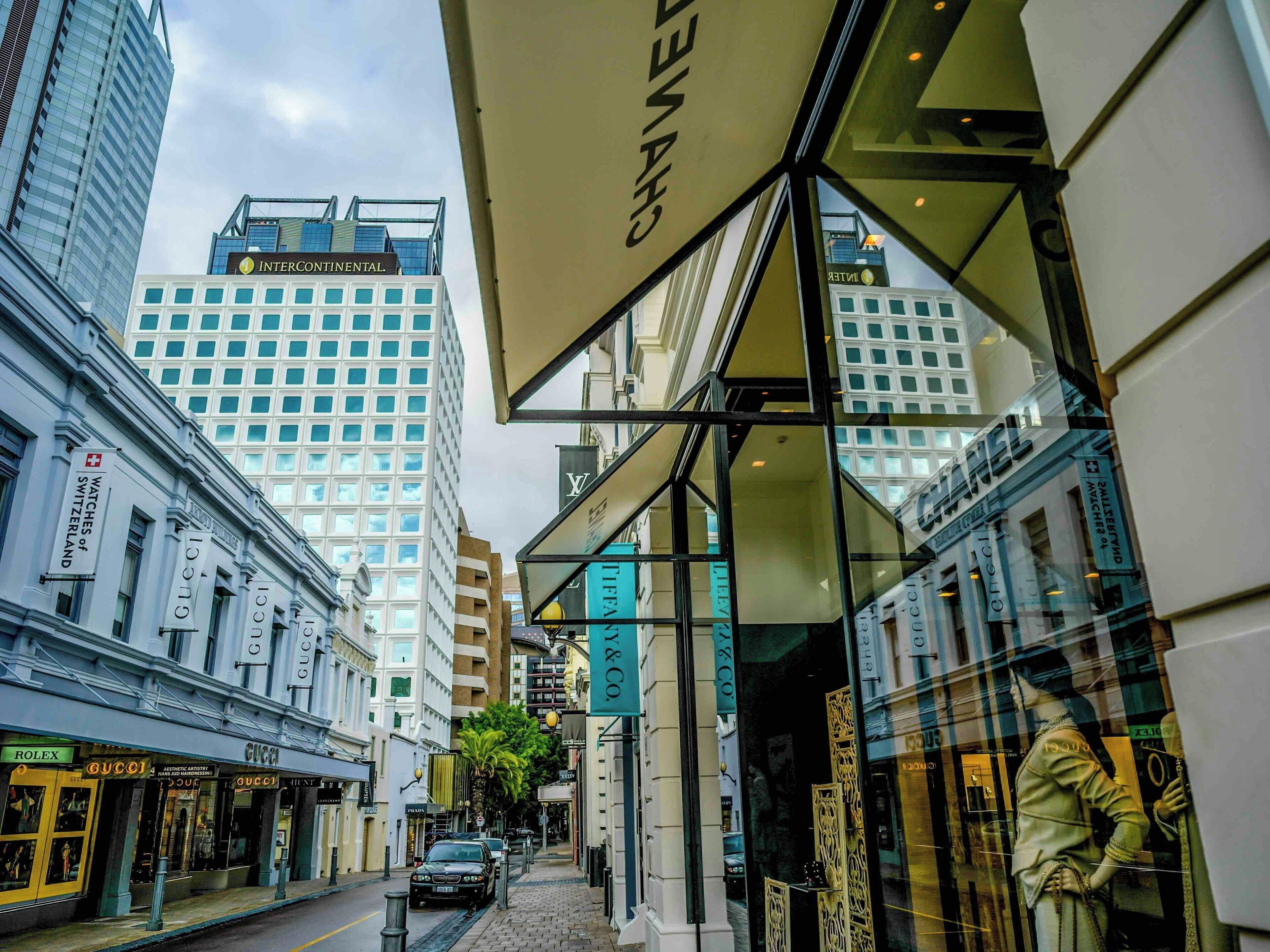 A street view with luxury brand storefronts, including Gucci and Rolex, and a high-rise building in the background.