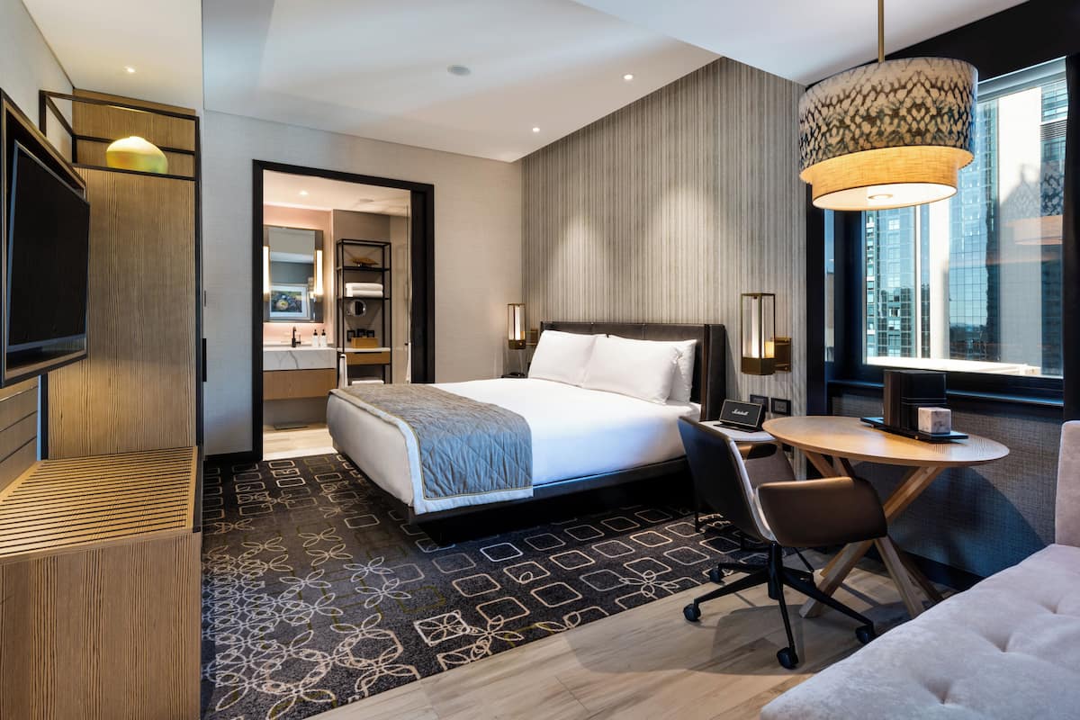 A modern hotel room with a large bed, a desk, a chair, a TV, a lamp, and a view of the city through the window.