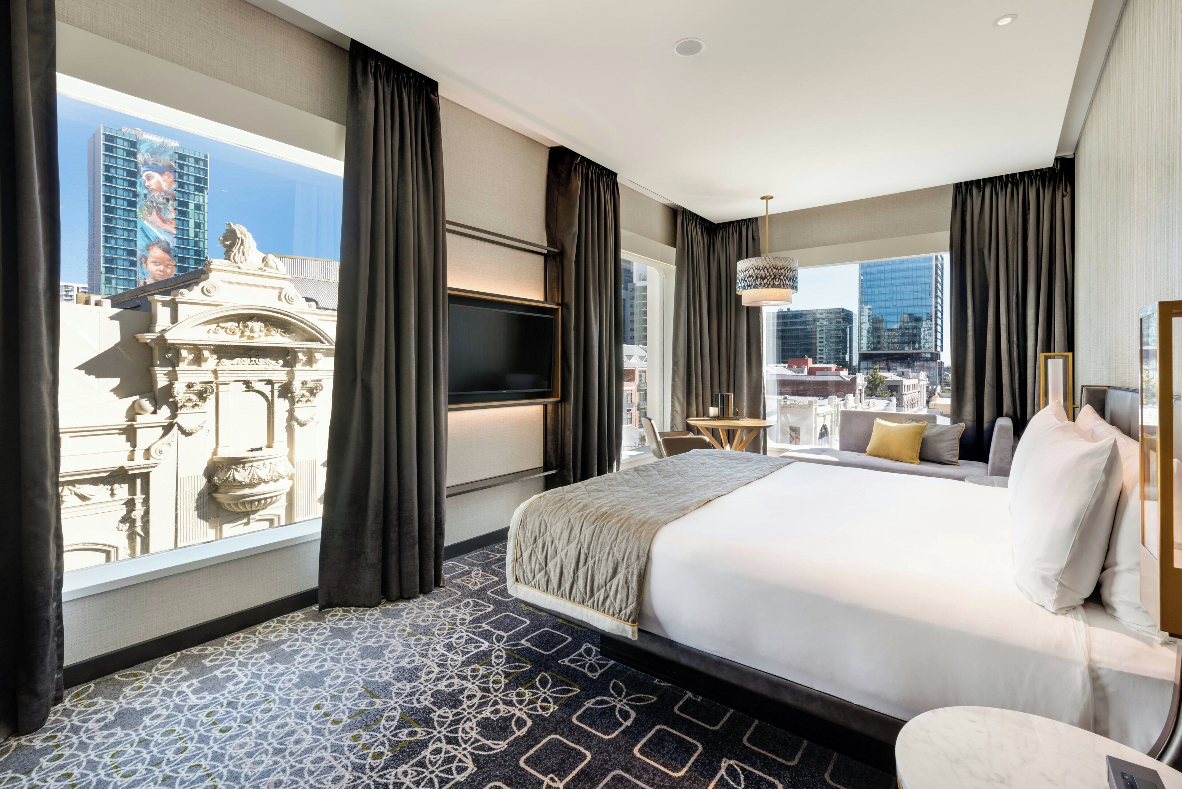 Ein Hotelzimmer mit einem großen Bett, einem Fernseher, Blick auf die Stadt und einem Balkon mit Tisch und Stühlen.
