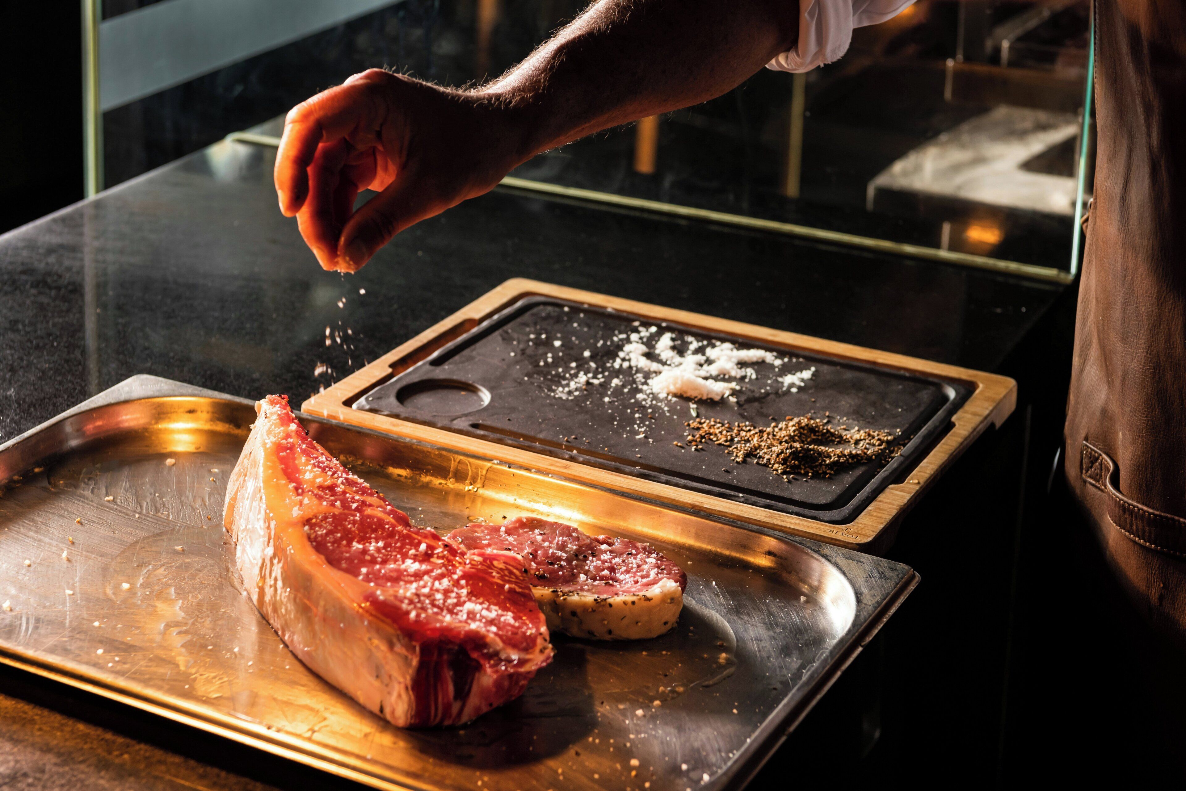 A chef seasoning a steak with various spices on a metal tray.