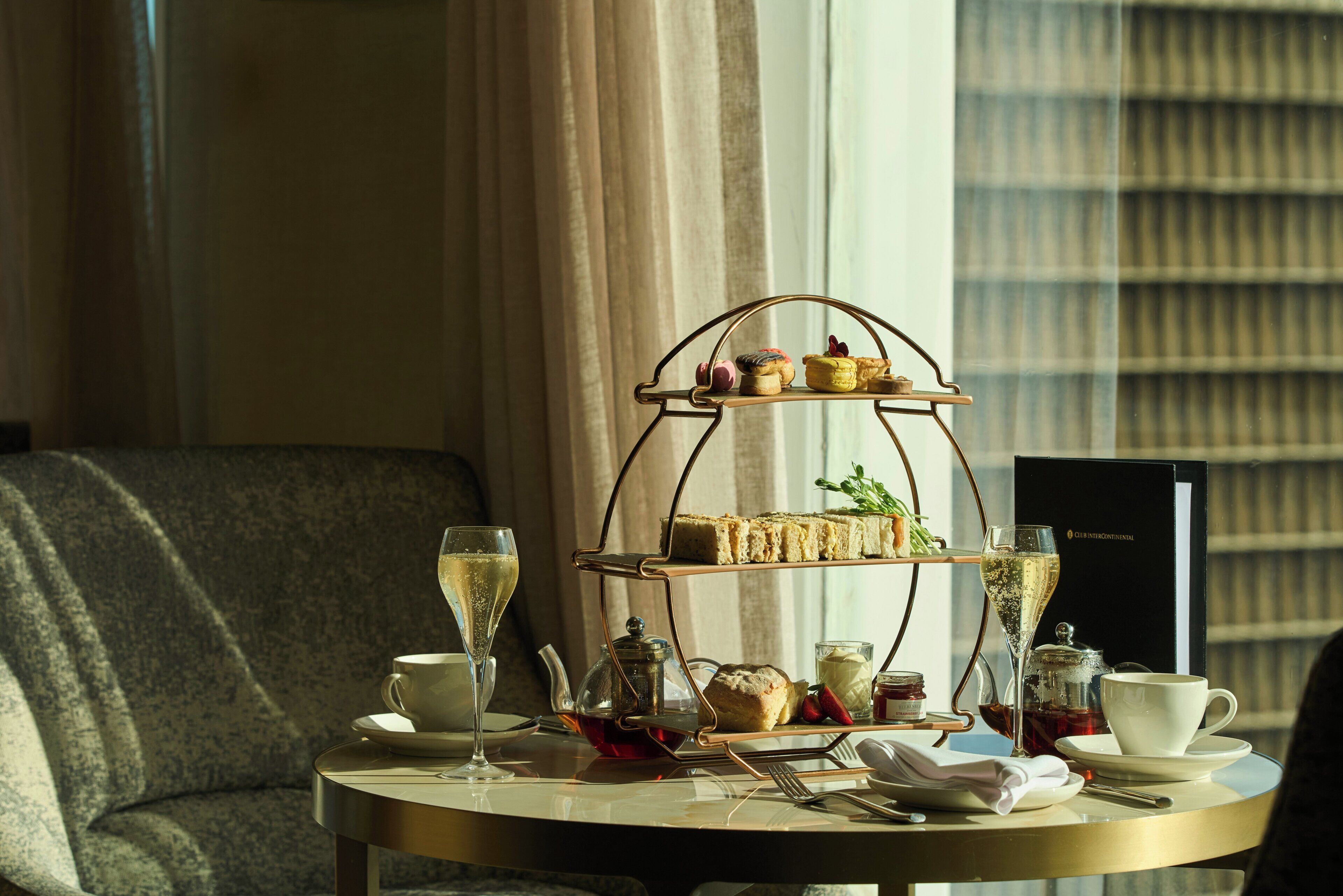 Un soporte de tres niveles con pasteles y sándwiches, acompañado de té y café, sobre una mesa redonda en una habitación con sofá y cortinas.