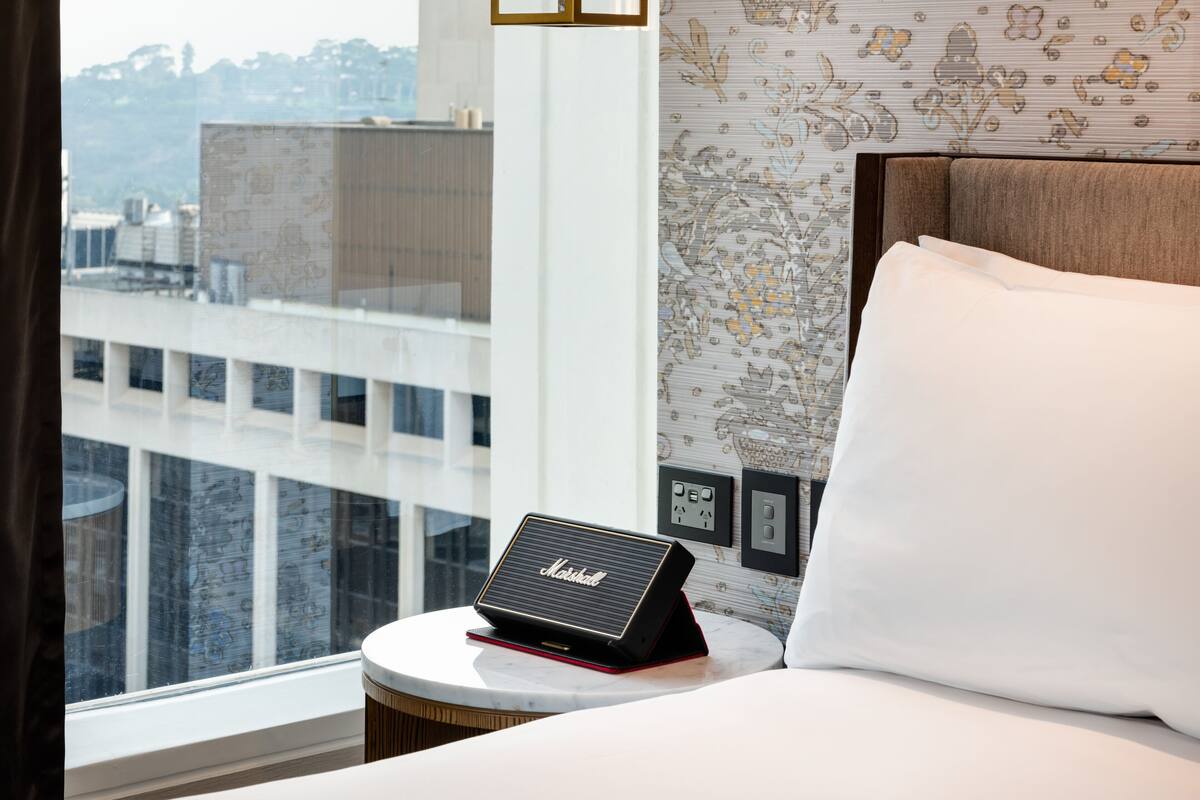 A hotel room with a bed, a small table with a speaker, a window with a view of buildings, and a patterned wall.