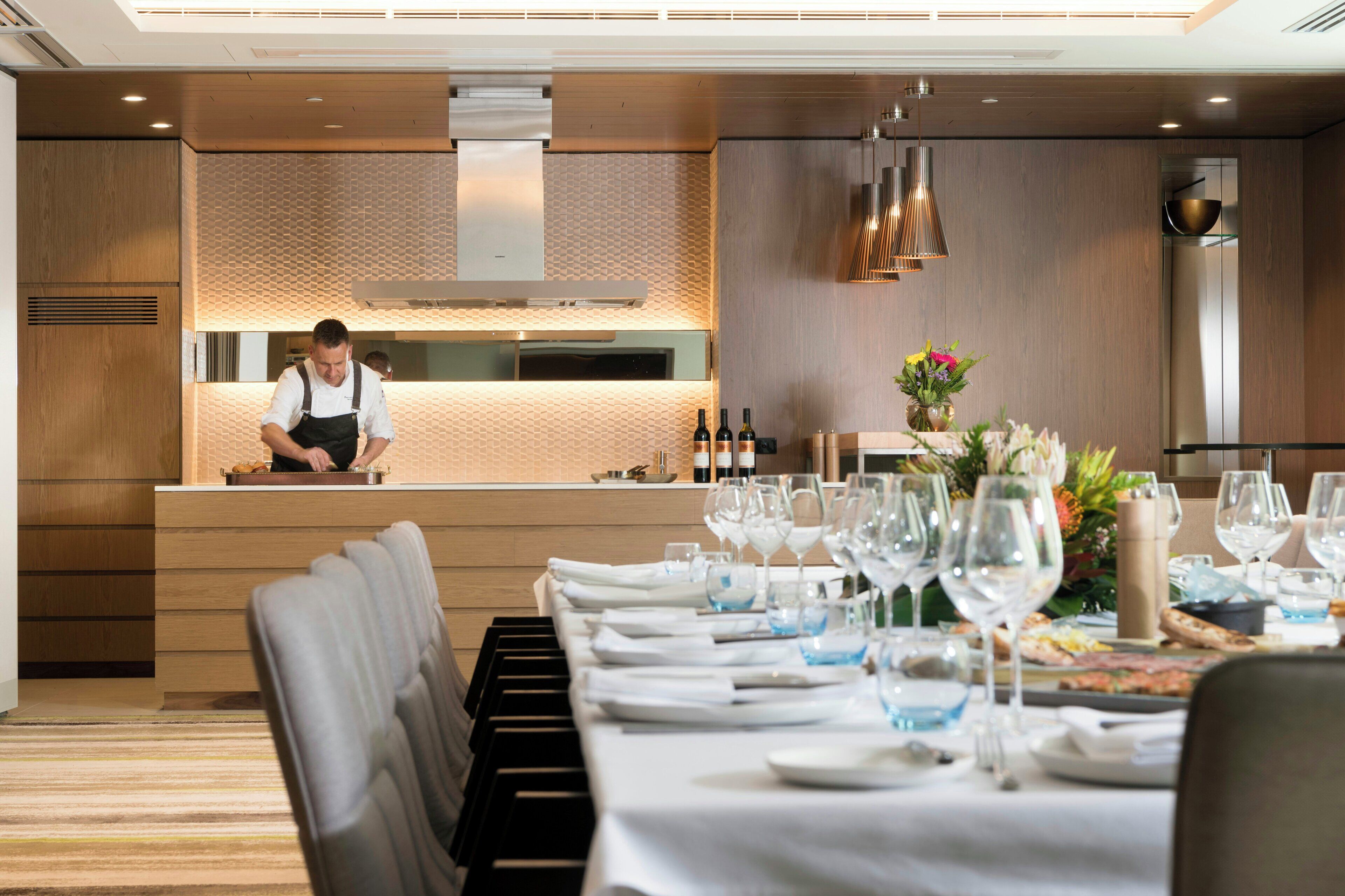 Un restaurante con un chef preparando comida, una larga mesa de comedor con mantel blanco, copas de vino y un jarrón con flores.