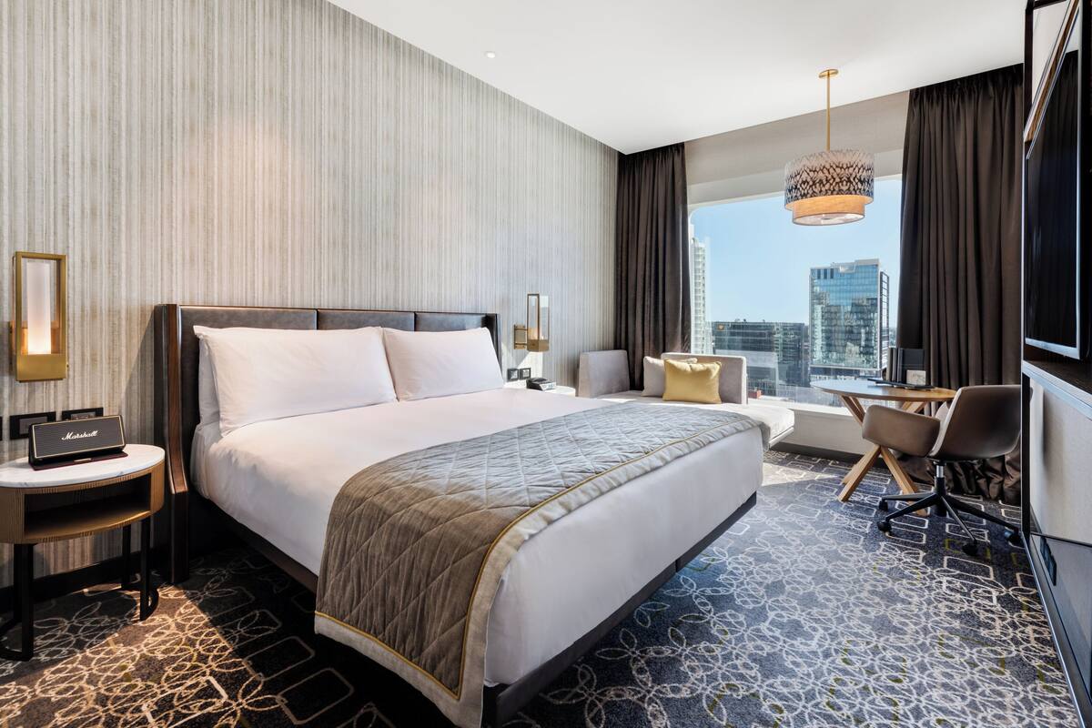 A modern hotel room with a large bed, a desk, a chair, and a view of the city skyline.