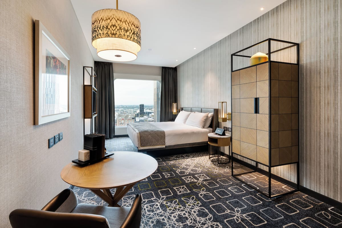 A modern hotel room with a large bed, a round dining table, a seating area, and a view of the city.