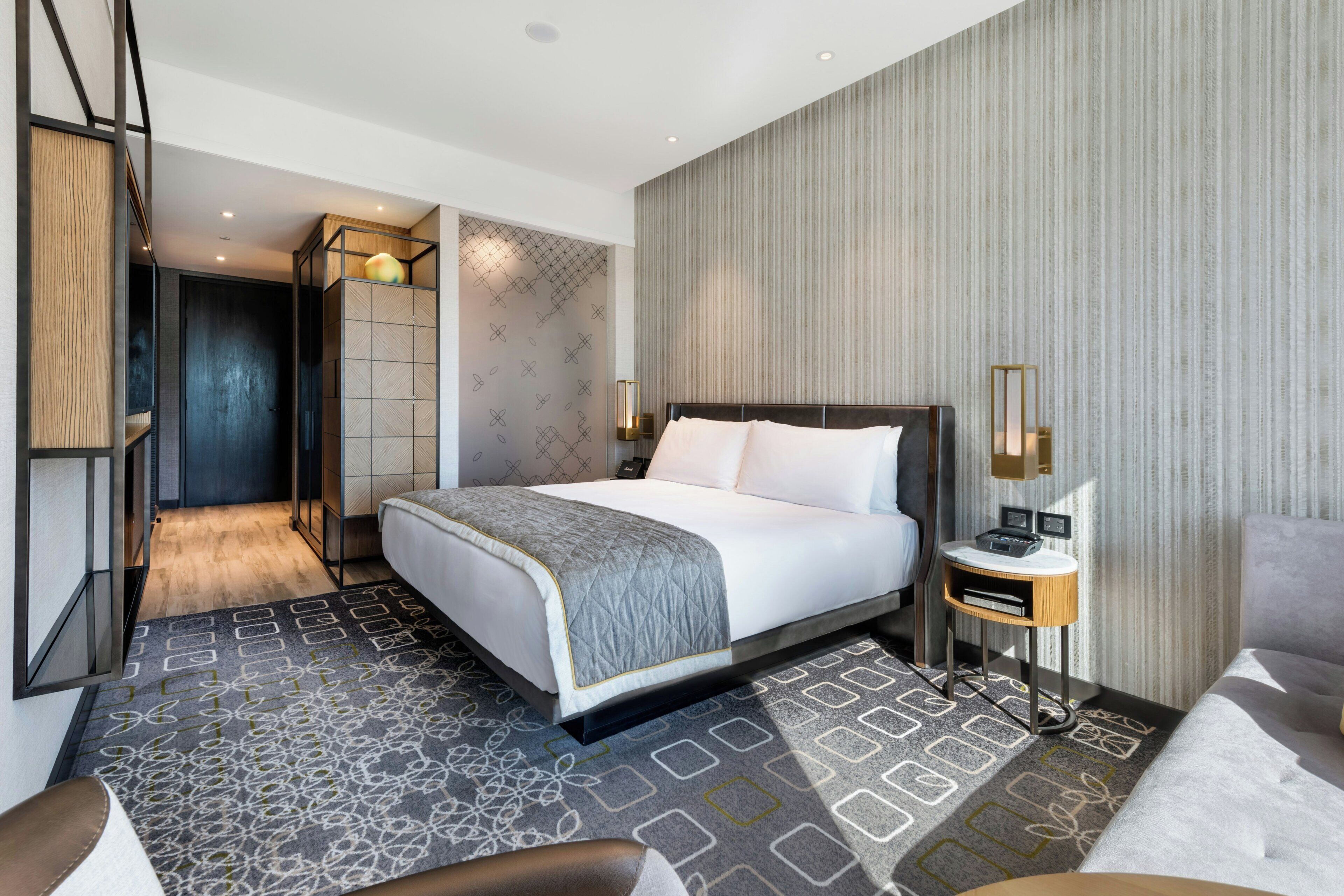 A modern hotel room with a large bed, a bedside table with a lamp, a seating area with a chair, and a view of the hallway.