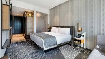 A modern hotel room with a large bed, a bedside table with a lamp, a seating area with a chair, and a view of the hallway.