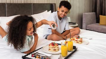 A hotel room with a bed, a bedside table, and a tray with breakfast items including pancakes, fruit, and juice.