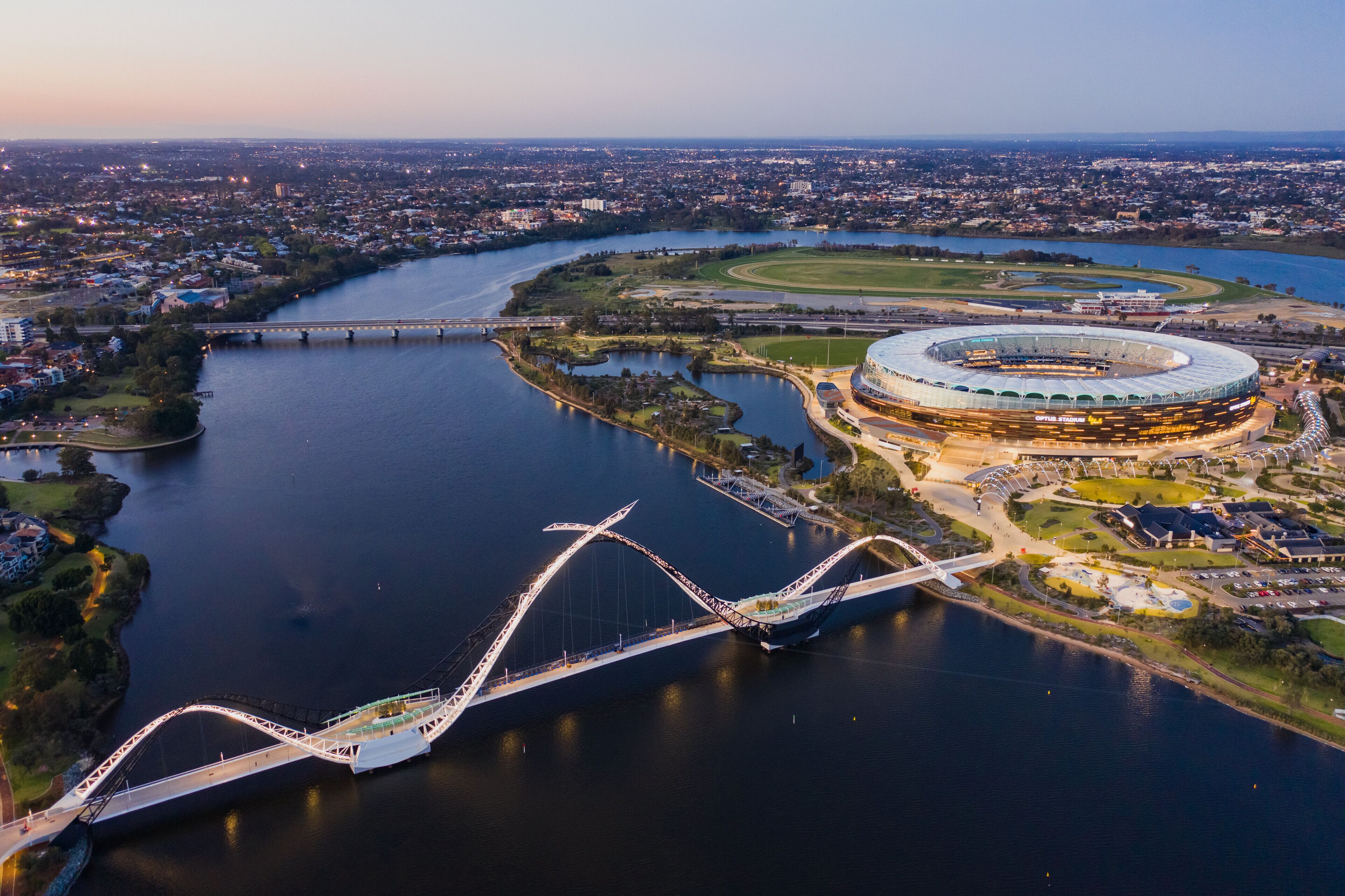An aerial view of a stadium with a circular design, surrounded by a river and a bridge.