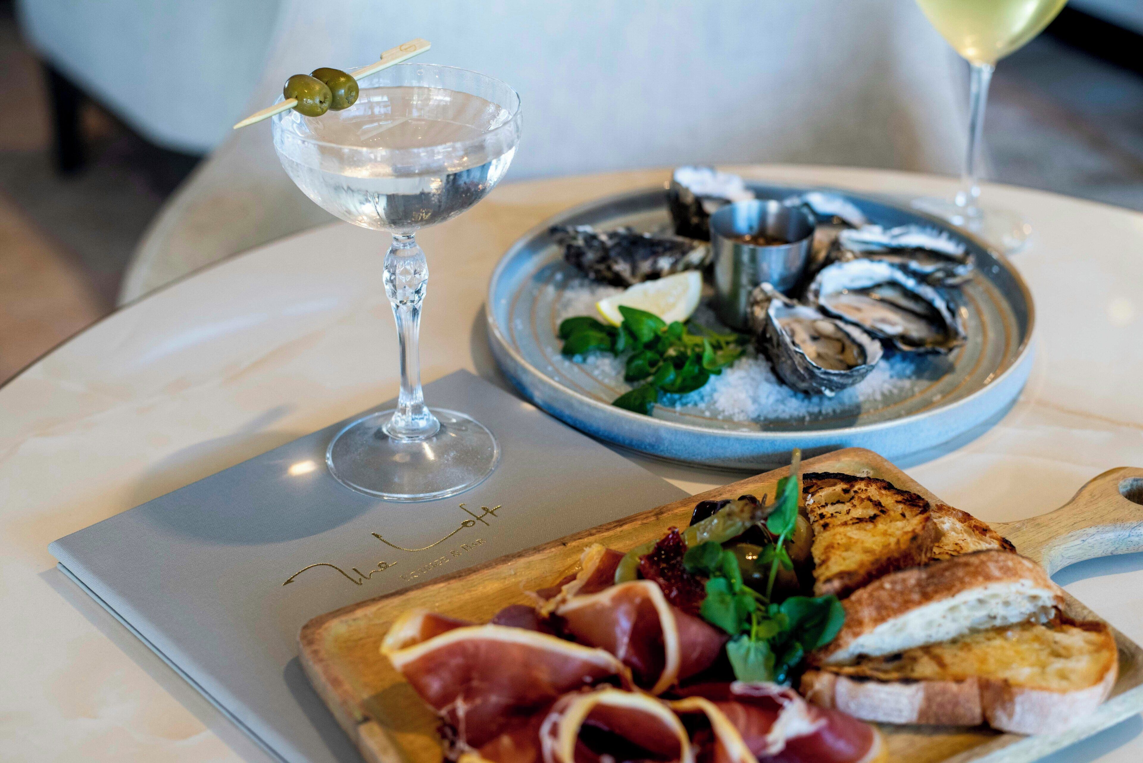 A dining table with a plate of oysters, a glass of white wine, and a sandwich with prosciutto and greens.