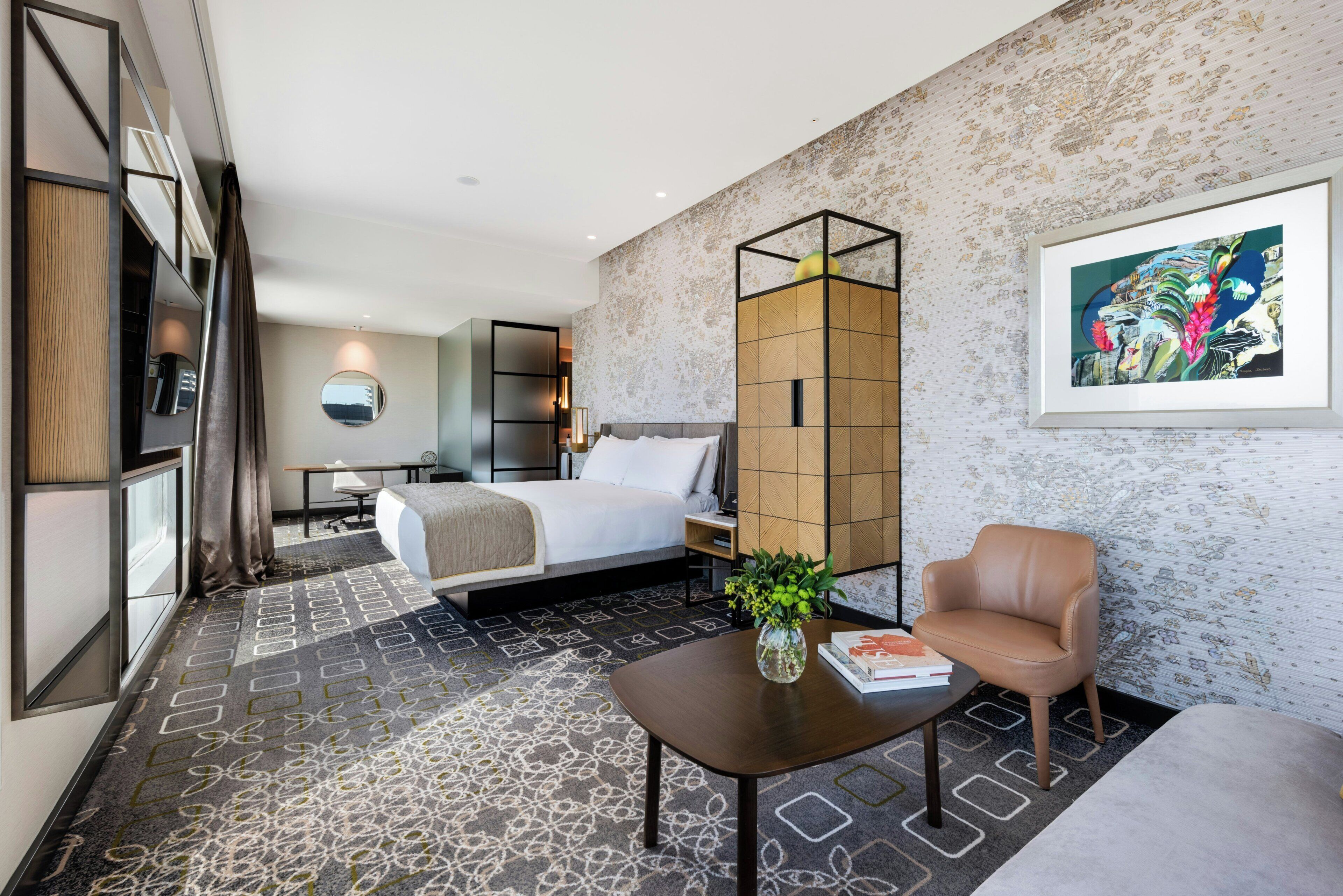 A modern hotel room with a bed, a desk, a chair, a sofa, a decorative vase, and a framed artwork on the wall.