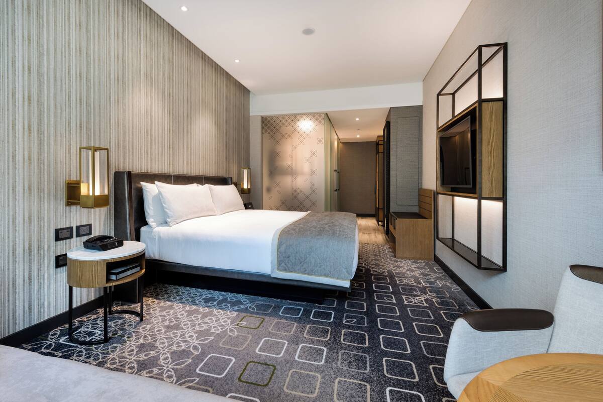 A modern hotel room with a large bed, a bedside table with a lamp, a TV mounted on the wall, and a patterned carpet.