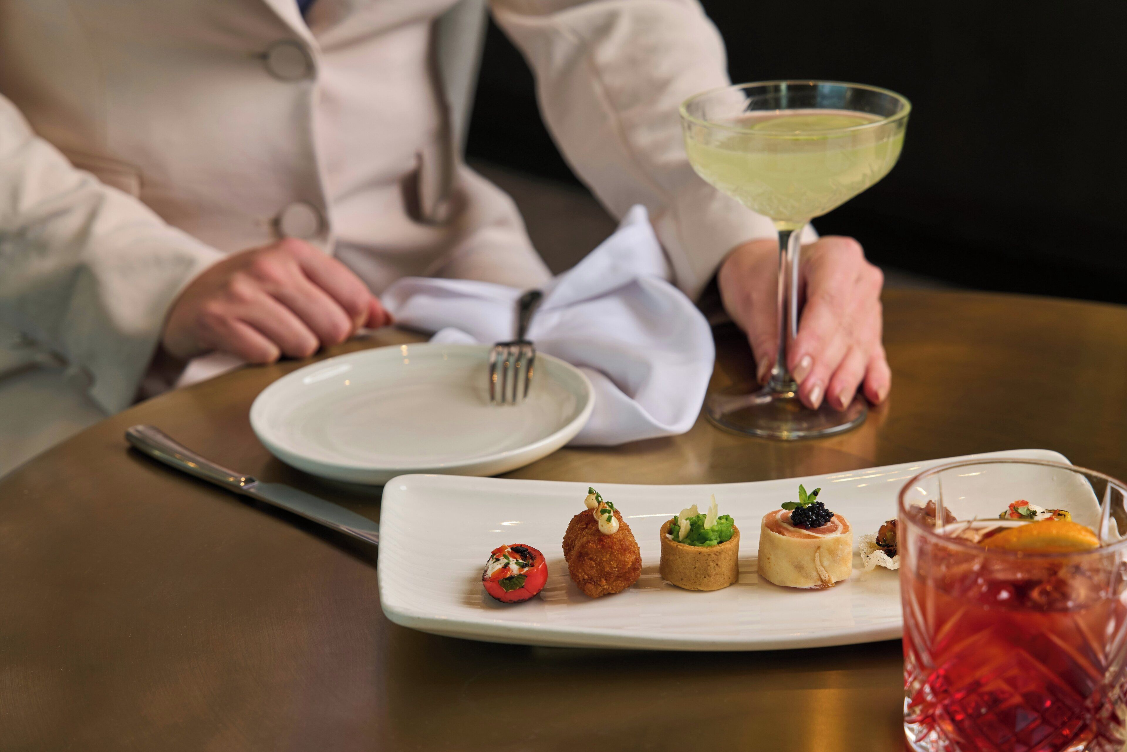 Una mesa con un plato blanco de aperitivos, un tenedor y un vaso de una bebida amarillenta.