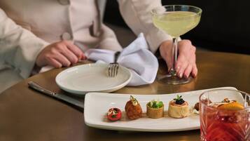 Una mesa con un plato blanco de aperitivos, un tenedor y un vaso de una bebida amarillenta.