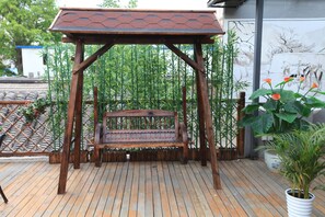 A wooden swing bench under a hexagonal roof, surrounded by bamboo plants and potted flowers.