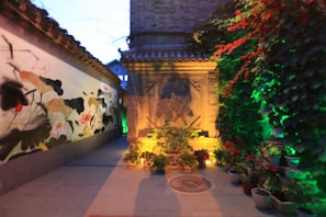 A narrow alley with a mural on the left, a traditional building with a tiled roof in the center, and potted plants on the right.