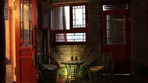 Una habitación con puerta roja, dos sillas de mimbre, una mesa pequeña y una ventana con cortinas.