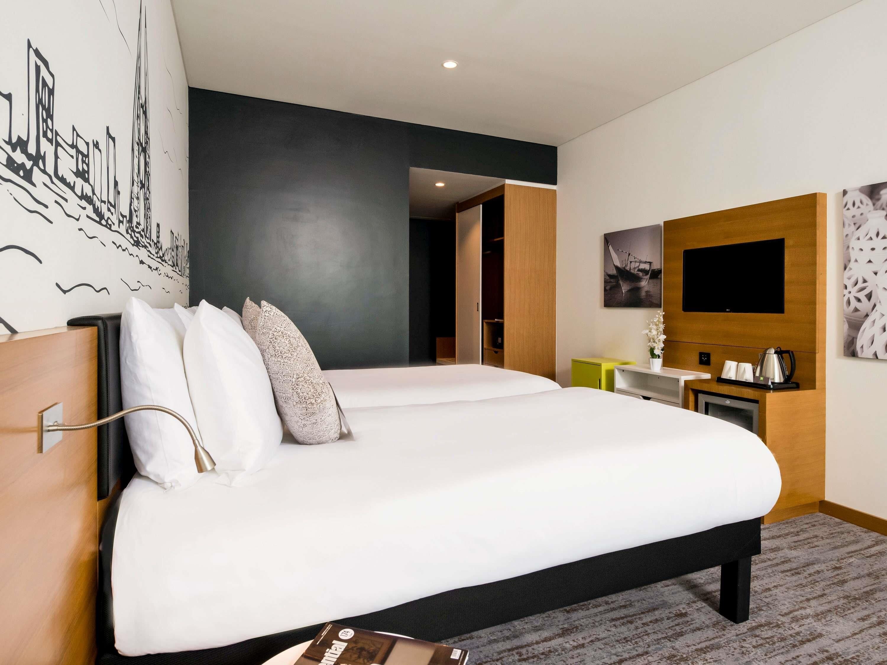 A modern hotel room with a large bed, a flat-screen TV, a small desk with a coffee maker, and a wall-mounted TV.