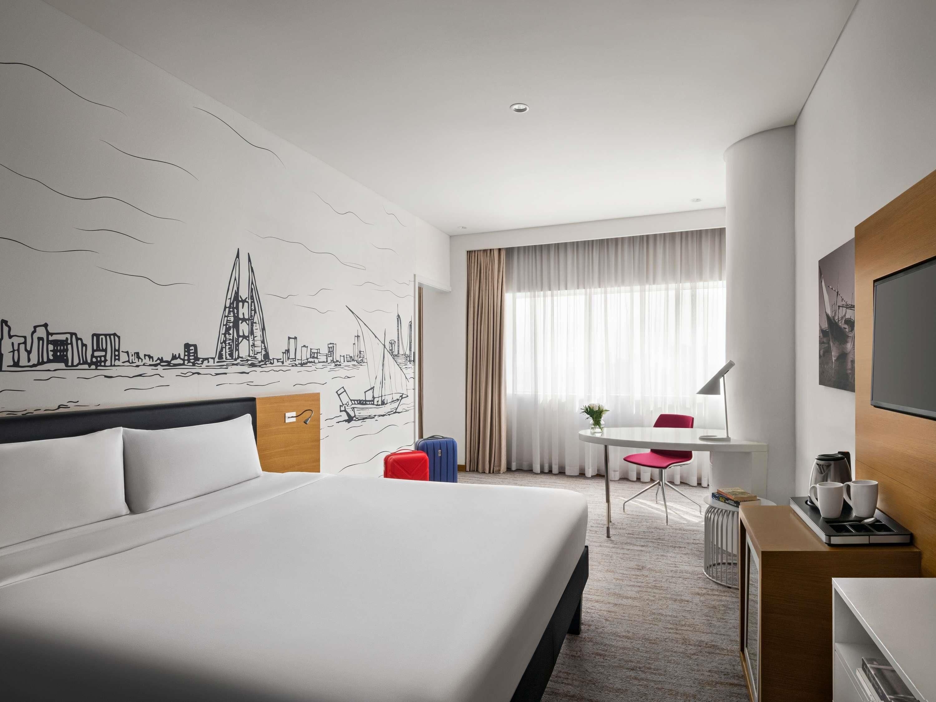 Habitación de hotel con una cama grande, un escritorio con silla, una pequeña mesa con otra silla, un televisor y un mural de paisaje urbano en la pared.