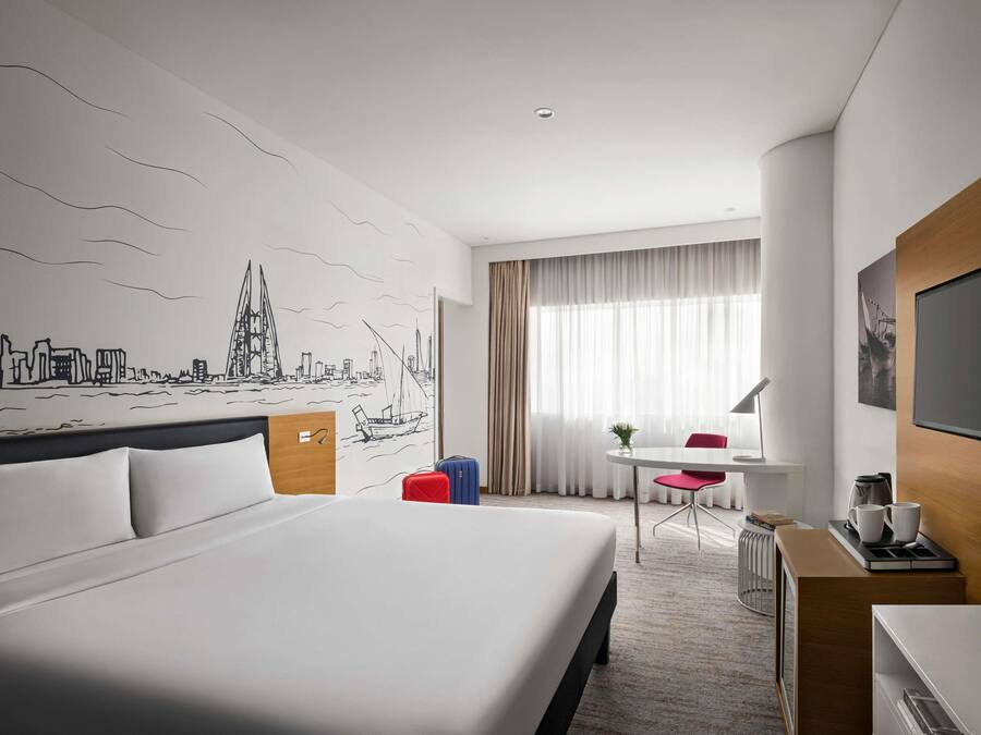 Habitación de hotel con una cama grande, un escritorio con silla, una pequeña mesa con otra silla, un televisor y un mural de paisaje urbano en la pared.