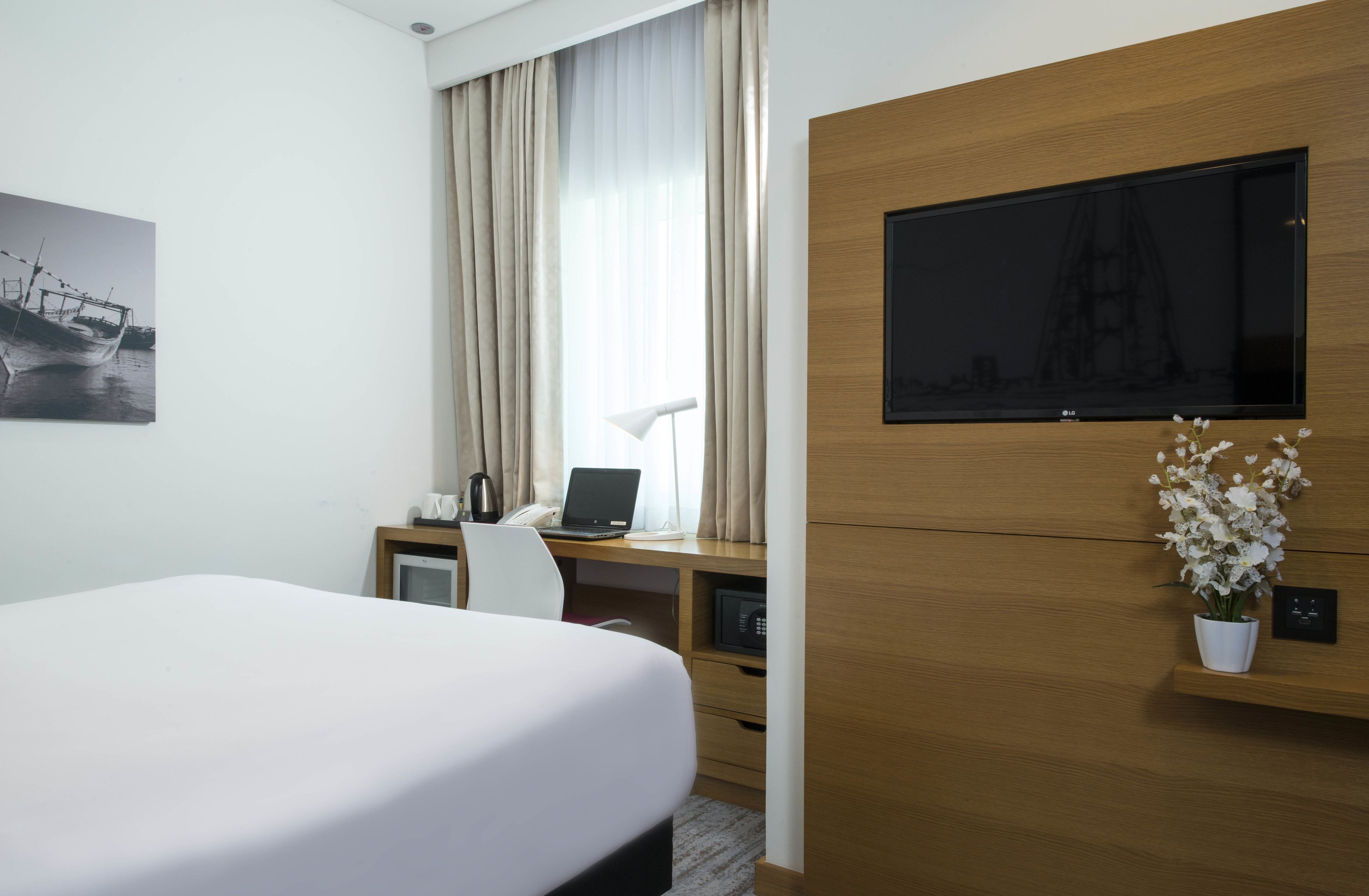 A hotel room with a bed, a desk with a laptop, a TV, and a small potted plant.