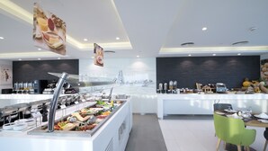 Un bufé de desayuno moderno con diversas opciones de comida, una estación de café y un área de estar con vistas al horizonte de la ciudad.