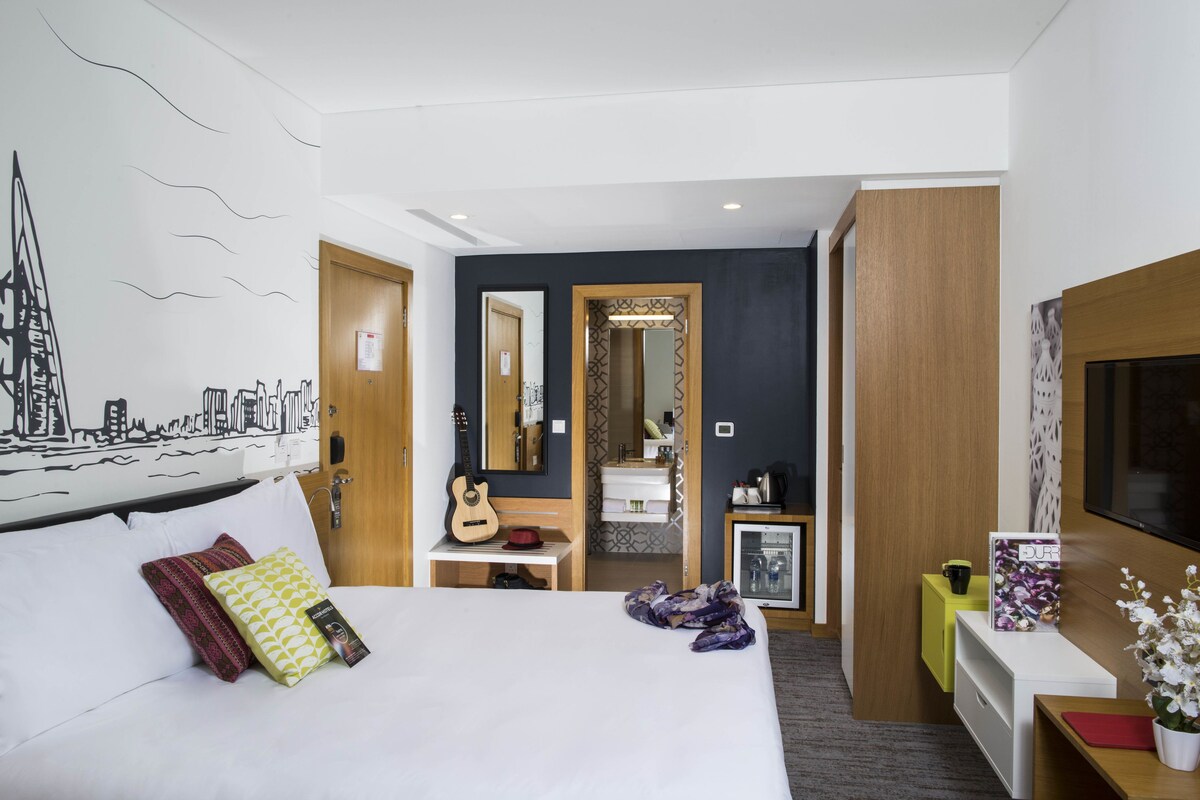 Una habitación de hotel moderna con una cama, una guitarra, un espejo, una tocador y un televisor.
