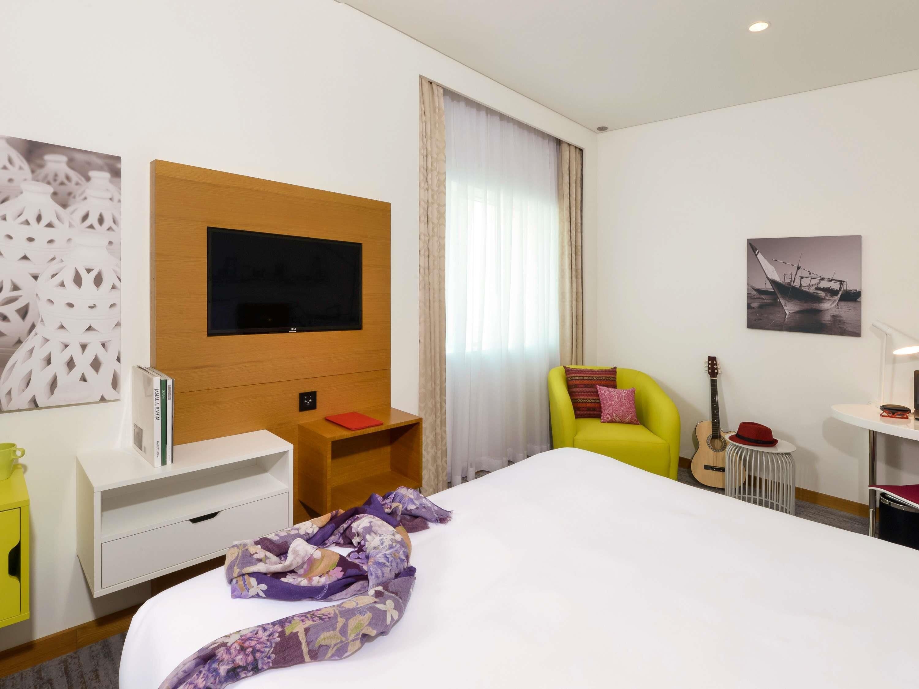 A hotel room with a bed, a TV mounted on a wooden panel, a chair, a guitar, and a small table with a lamp.