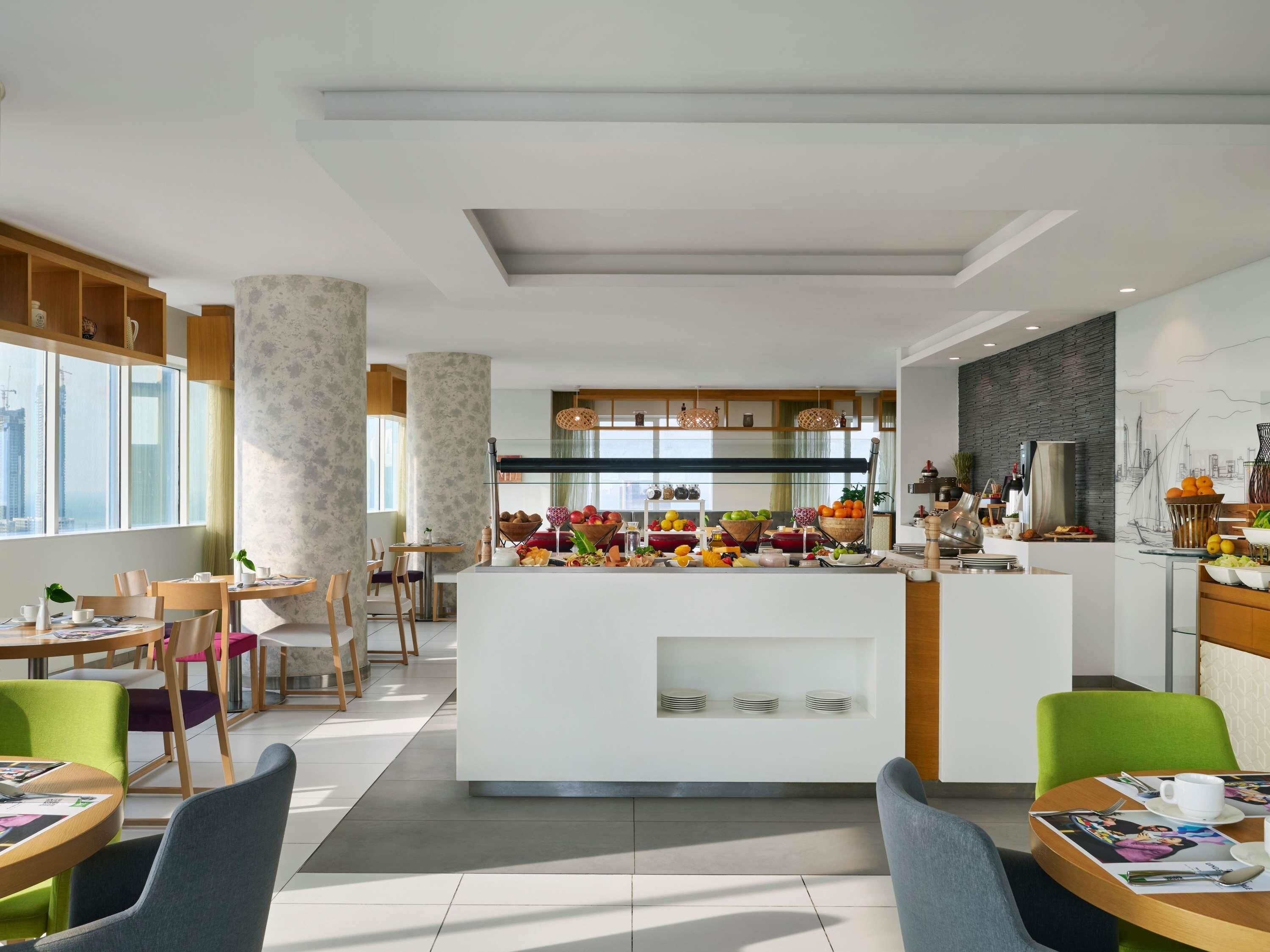 Een moderne hotel lobby met een centraal eiland, een schaal met fruit, zithoeken met stoelen en tafels, en een uitzicht op de stadshemel door grote ramen.