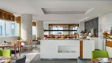 Een moderne hotel lobby met een centraal eiland, een schaal met fruit, zithoeken met stoelen en tafels, en een uitzicht op de stadshemel door grote ramen.
