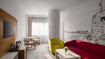 Una habitación de hotel moderna con un televisor de pantalla plana, una zona de comedor con mesa y sillas, un sofá rojo vibrante, un sillón amarillo y un mural con un paisaje urbano en la pared.